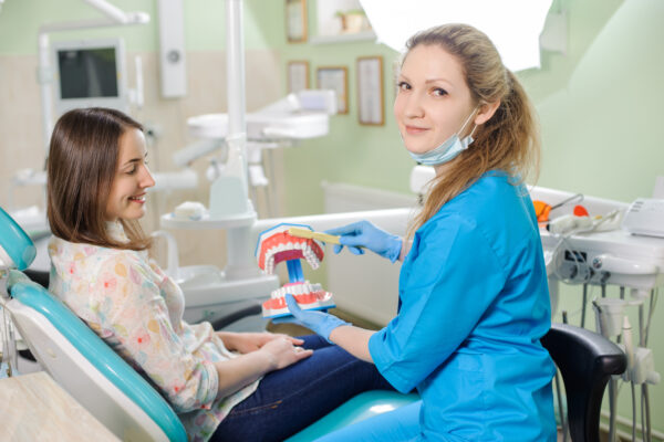 Young female dentist showing dental jaw model to woman patient in dentist's clinic. Smiling woman at dentist sitting in dentist chair. Dentistry