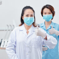 Portrait of cheerful experienced dentist and nurse in protective masks and silicone gloves showing thumbs-up and looking at camera