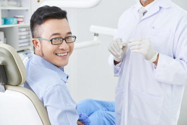 Horizontal shot of cheerful Asian patient of dental clinic sitting on chair smiling, unrecognizable dentist standing next to him