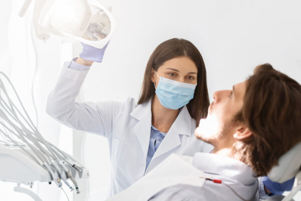 Dentist doctor woman in mask turning on lamp before making check up, patient sitting with open mouth