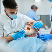 Young dentist polishing teeth of senior patient during dental procedure at clinic. Focus is on senior man.